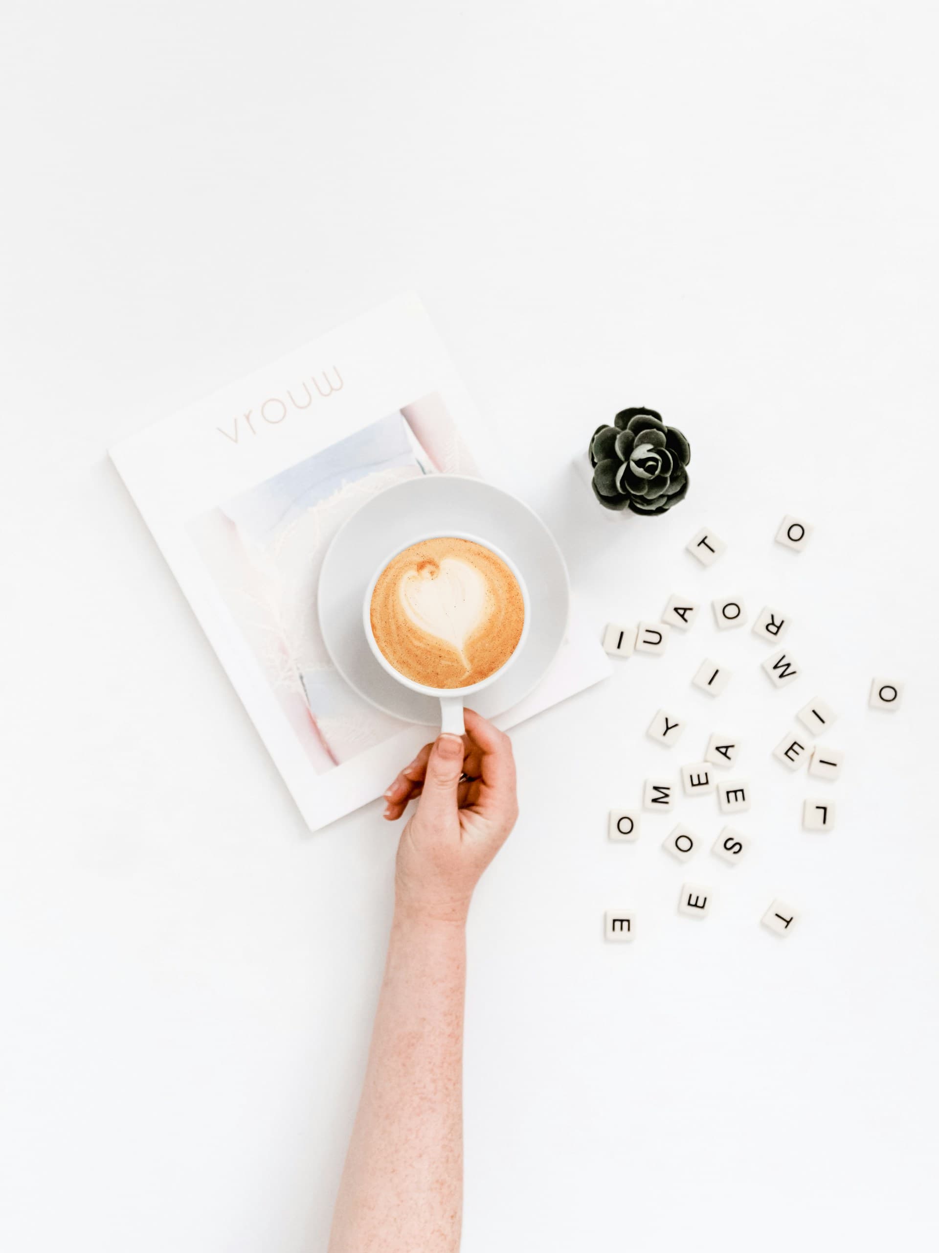 Coffee cup held over a magazine by a hand with Scrabble letters scattered to its right.