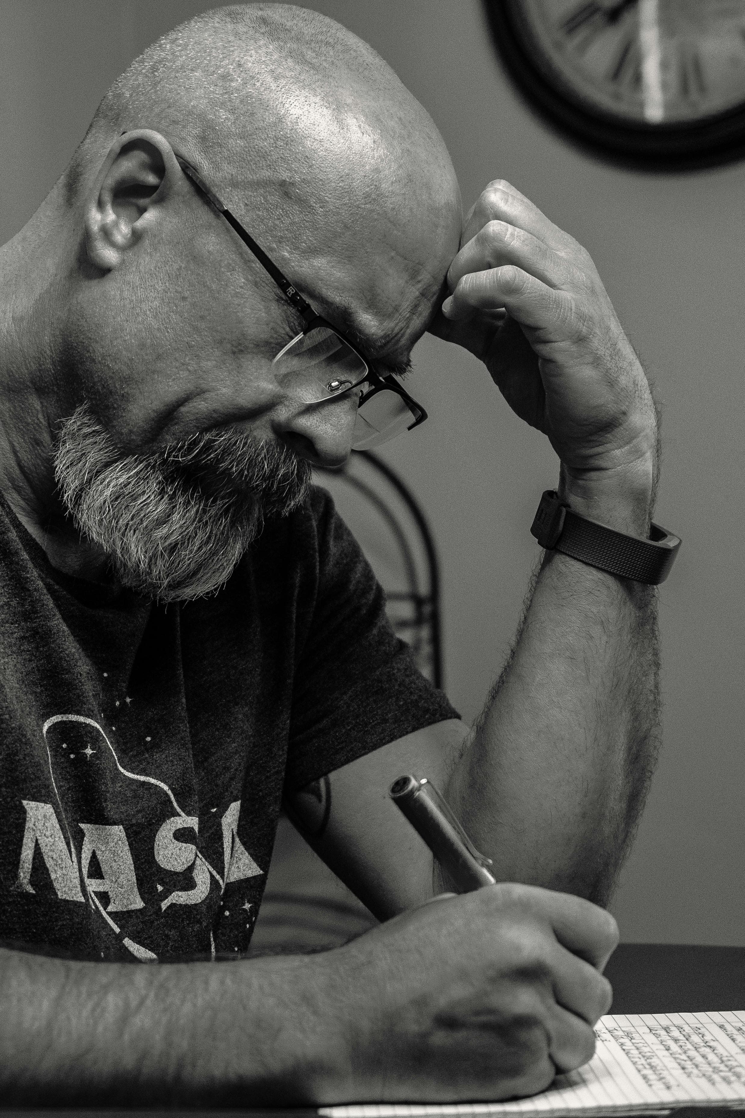 Bald man with beard and glasses in a NASA shirt concentrates on writing.